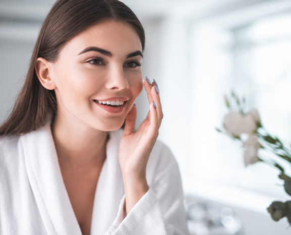 Portrait of pleased young lady touching cheek with hand. She is putting cream on face indoors