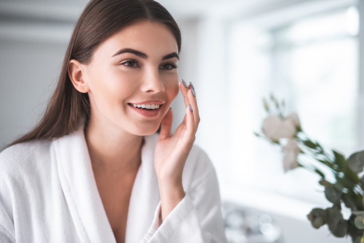 Portrait of pleased young lady touching cheek with hand. She is putting cream on face indoors