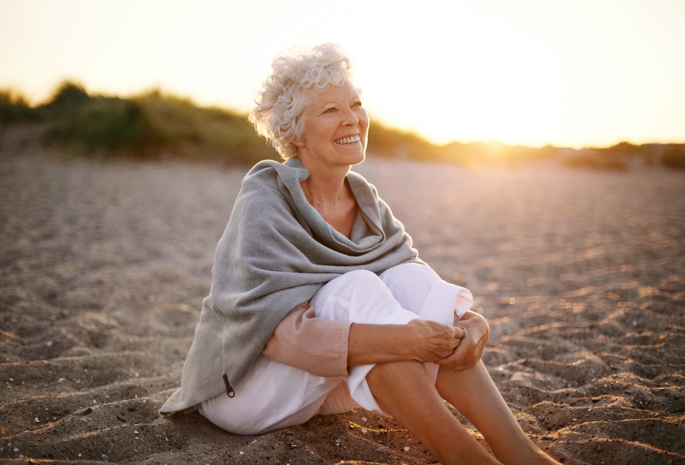An older woman wearing white cropped pants and a knitted poncho sites on the beach at sunrise and smiles